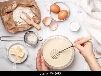 La donna si prepara la pasta per frittelle fatte in casa per la prima colazione. Mescolare con una frusta per la fustigazione in mani. Gli ingredienti sul tavolo - farina di frumento, uova, burro, zucchero, sa Foto Stock