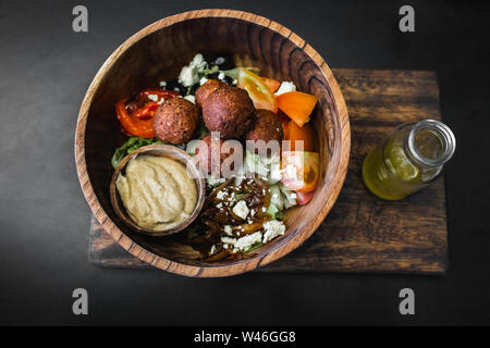 Ciotola di legno con sfere di falafel, hummus e insalata fresca. Olio d'oliva in bottiglia sul lato. Dark sfondo nero, vista dall'alto. Tradizionale cibo israeliano. Vegani Foto Stock