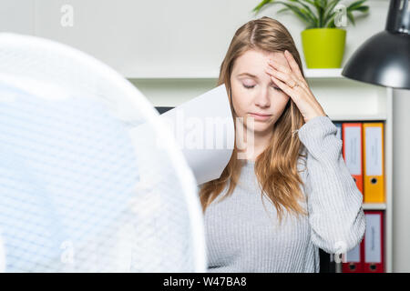 Una donna in ufficio soffre dal calore e utilizzando un ventilatore per il raffreddamento Foto Stock