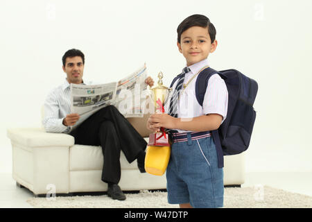 Scolaro che tiene un trofeo con suo padre che legge giornale dentro lo sfondo Foto Stock