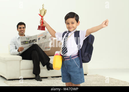 Scolaro che tiene un trofeo con suo padre che legge giornale dentro lo sfondo Foto Stock