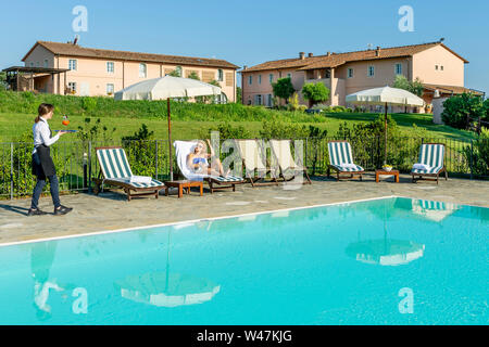 Giovane cameriera serve un cocktail a bordo piscina per un cliente seduto su una sedia a sdraio in un resort nel contado di Pisa, Toscana, Italia Foto Stock