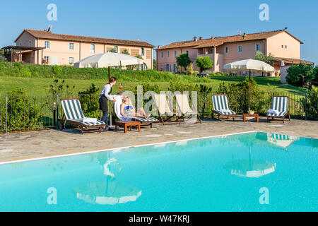 Giovane cameriera serve un cocktail a bordo piscina per un cliente seduto su una sedia a sdraio in un resort nel contado di Pisa, Toscana, Italia Foto Stock