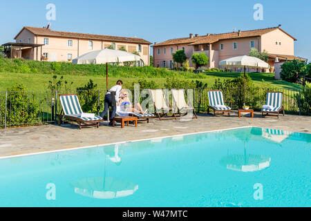 Giovane cameriera serve un cocktail a bordo piscina per un cliente seduto su una sedia a sdraio in un resort nel contado di Pisa, Toscana, Italia Foto Stock