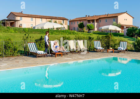 Giovane cameriera serve un cocktail a bordo piscina per un cliente seduto su una sedia a sdraio in un resort nel contado di Pisa, Toscana, Italia Foto Stock