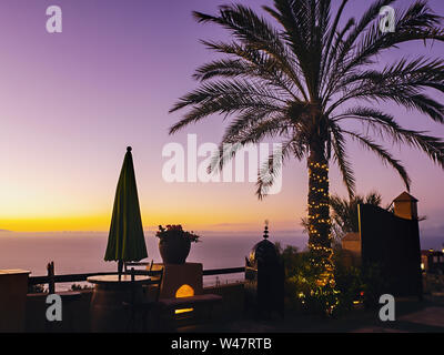 Viola golden atmosfera serale sulla costa nord di Tenerife e con una grande palma circondata dalla catena di luce su una visualizzazione di terrazza che si affaccia sul Foto Stock