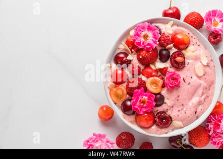 Ciotola frullato o Nizza crema fatta di banane surgelate e frutti di bosco con frutti di bosco freschi, rose fiori, noci e semi su marmo bianco tavolo dove siamo sani Foto Stock