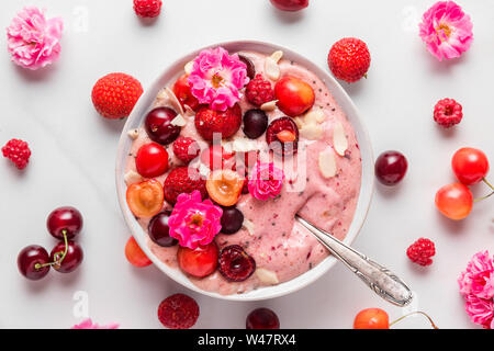 Crema di Nizza o frappè ciotola fatta di banane surgelate e bacche con rose fiori, frutta a guscio e i semi con un cucchiaio su marmo bianco tavola. una sana prima colazione Foto Stock