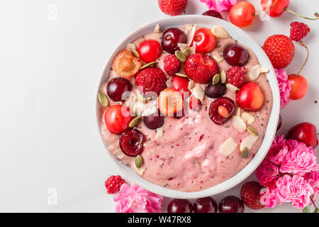 Dieta sana prima colazione. smoothie ciotola o Nizza crema fatta di banane surgelate e frutti di bosco con frutti di bosco freschi, rose fiori, noci e semi su bianco marbl Foto Stock