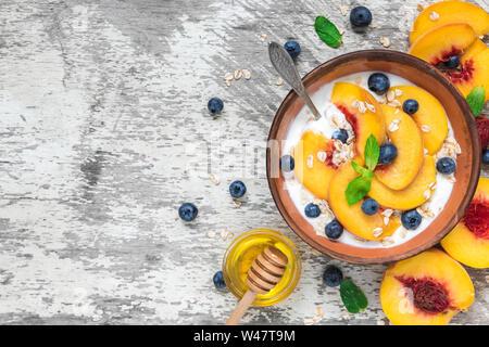 Ciotola di yogurt greco con pesca, avena, dadi, menta, miele e mirtilli con un cucchiaio per una sana prima colazione. vista superiore Foto Stock