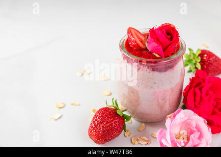 Romantica Colazione sana. Pernottamento strawberry avena con frutti di bosco freschi e menta con rose fiori in un bicchiere su marmo bianco tavola. close up Foto Stock