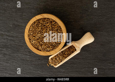 Sacco di tutta la secca di frutti di cumino in una ciotola di legno con il cucchiaio di legno flatlay sulla pietra grigia Foto Stock