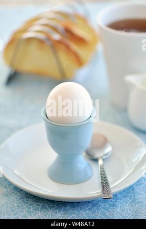 Bollito di fattoria fresco bianco uovo in eggcup blu su bianco e blu tovaglia modellato con pane tostato e tè. In formato verticale con il fuoco selettivo su eggc Foto Stock