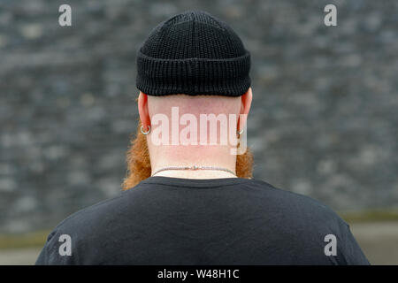 Vista posteriore della testa rasata di un uomo con sun bruciato collo indossando un Beanie black hat, orecchini e catena argento Foto Stock