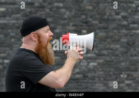 Arrabbiato barbuto man in black beanie hat gridando in un megafono arieggiare le sue lamentele in corrispondenza di uno sciopero o di dimostrazione su uno sfondo grigio Foto Stock