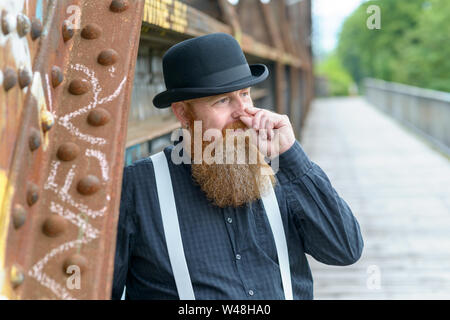 Uomo Barbuto in bombetta all'aperto permanente strofinando il naso come egli si appoggia contro un vecchio arrugginito supporto metallico di un ponte Foto Stock