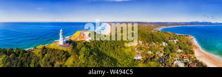 In alto di capezzagna con Byron Bay lighthouse alta sopra la costa dell'oceano Pacifico in una giornata di sole in elevata panorama antenna rivolta verso l'interno sopra le spiagge sabbiose. Foto Stock