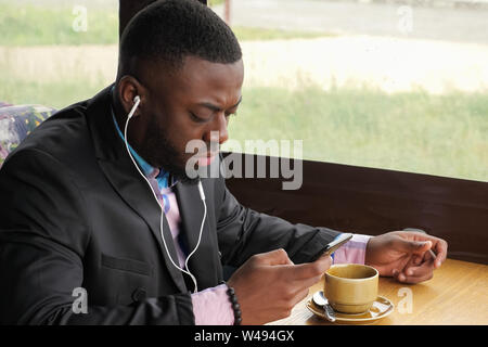 Afro American imprenditore nel cafe. Uomo nero è in ascolto di musica in cuffia la navigazione del telefono cellulare e di bere il caffè in seduta cafe. Ragazzo indossa in maglietta e tuta giacca. Godendo di mattina tempo. Foto Stock