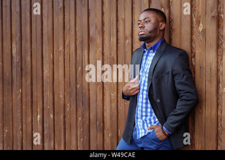 Ritratto di giovane nero barbuto guy standing e la mano in tasca. Afro-americano in tuta è fiducioso e guarda lontano su legno scuro sfondo, copyspace Foto Stock