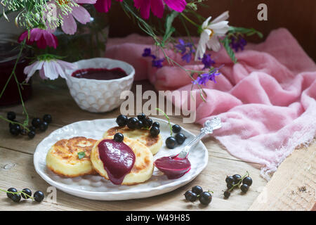 Colazione a base di frittelle di quark, ribes crema pasticcera, le uve secche di Corinto e un mazzo di fiori selvatici su uno sfondo di legno. Foto Stock