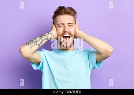 Nervoso frustrato infastiditi giovane con ampia bocca aperta la chiusura di orecchie con palme. Isolato su sfondo blu. uomo odia la musica ad alto volume, guy essendo infastidire Foto Stock