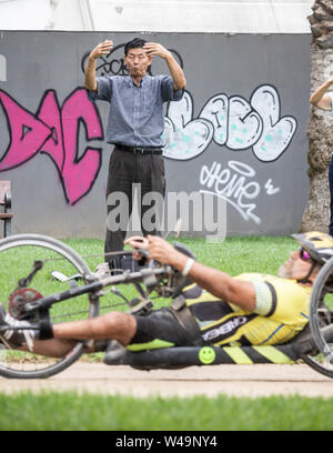 A Las Palmas di Gran Canaria Isole Canarie Spagna, 21 luglio, 2019. Piacere e dolore. Un concorrente disabili utilizzando una handbike passa su un gruppo di persone che praticano il Tai Chi durante l'ultima ora di una 24 ore di gara per il relè i team e i singoli in un 1km del circuito a Las Palmas de Gran Canaria. Credit:Alan Dawson/Alamy Live News Foto Stock