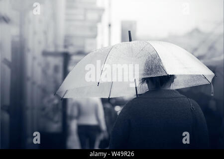 Una ragazza solitaria cammina lungo una strada di città in foggy meteo sotto un ombrello trasparente che protegge il suo dalla pioggia. In bianco e nero il colore aggiunge un sa Foto Stock