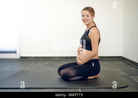 Gravidanza felice giovane donna facendo esercizi inginocchiati su un materassino yoga stringendo la sua pancia gonfia in entrambe le mani mentre sorridente verso la telecamera Foto Stock