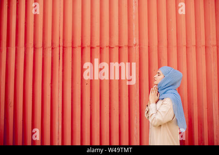 Giovane donna islamica in informale e hijab pregando contro la parete rossa Foto Stock