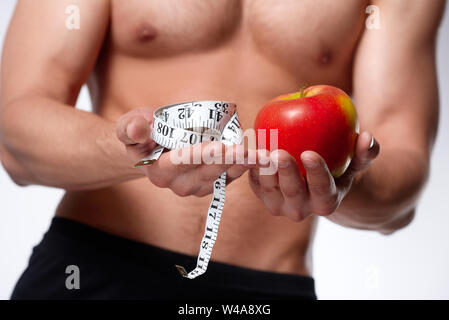 Uomo Forte con corpo muscoloso detiene il nastro di misurazione e red apple nelle sue mani. Concetto di sport e sana alimentazione. Isolato su sfondo bianco Foto Stock