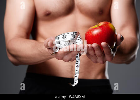 Uomo Forte con corpo muscoloso detiene il nastro di misurazione e red apple nelle sue mani. Concetto di sport e sana alimentazione. Isolato su sfondo grigio Foto Stock