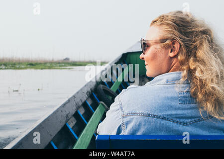 Caucasian donna bionda in denim shirt avente un giro in barca lungo i giardini galleggianti sul Lago Inle, Myanmar Foto Stock