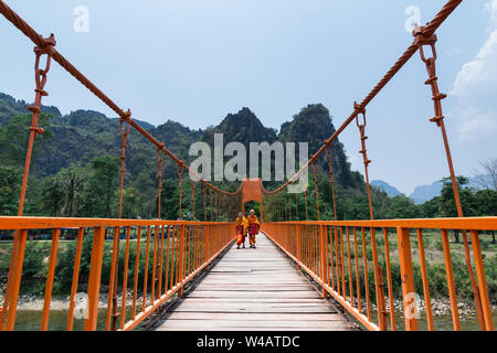 Vang Vieng, Laos - Aprile 2019: due monaci buddisti in arancione vesti attraversando ponte di sospensione. Foto Stock