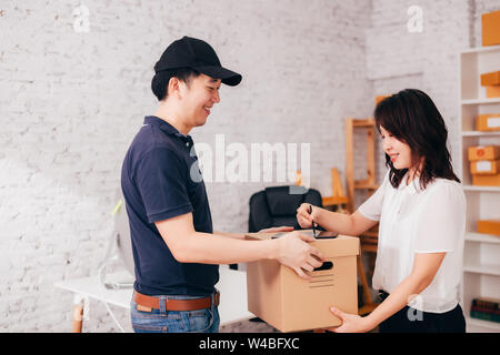 Vista laterale di sorridere Asian femmina tablet di firma durante l'assunzione di scatola di cartone da felice uomo consegna in ufficio Foto Stock