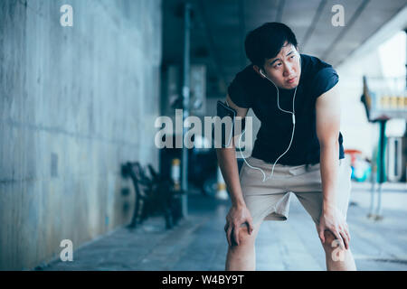 Stanco Asian guy nelle cuffie appoggiato sulle ginocchia e guardando lontano mentre ascolti la musica e rilassante durante l'allenamento all'aperto sulla strada di città Foto Stock