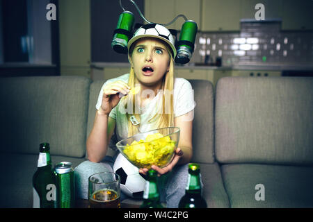 Giovane donna guardare la partita di calcio in tv di notte. Preoccupatevi model look up e mangiare patatine. Modello bionda indossare il cappello con due lattine di birra sul lato di essa. Bottiglia di un Foto Stock