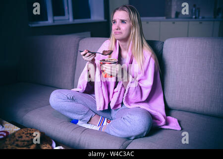 Giovane donna guardando film di notte. Seduto con le mani incrociate e mangiare dolci. I cookie sul tavolo. Emotivo donna preoccupati da soli in camera Foto Stock