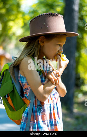 Ragazza bionda in un cappello con capelli lunghi passeggiate attraverso il parco con lo zaino sulle spalle e si mangia una mela su una soleggiata giornata estiva Foto Stock