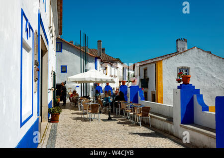 Scena di strada, ristorante esterno, tavoli, sedie e vecchi edifici di colore bianco e luminoso rivestimento blu, ciottoli, Europa Obidos, Portogallo, molla, orizzontale Foto Stock