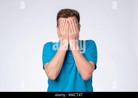 Giovane uomo caucasico in maglietta blu chiude il viso con le mani per cercare di restare anonym. Foto Stock