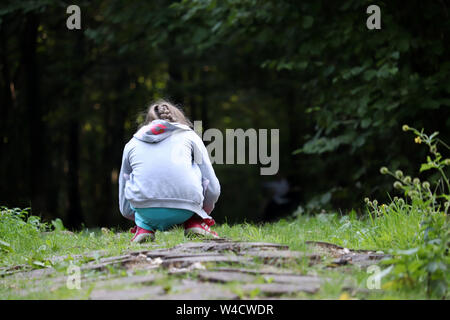 Bambina gioca sul percorso in una foresta scura, in vista posteriore Bambino squatting raccoglie le piante in estate Foto Stock