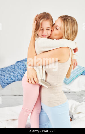 Con amore di madre abbraccia la figlia e dà un bacio a letto in camera da letto Foto Stock