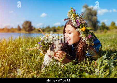 Pug cane e il suo padrone la refrigerazione dal lago indossando ghirlande di fiori. Happy puppy leccare la donna in estate park Foto Stock