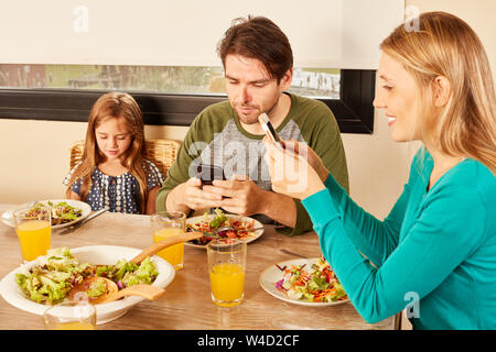 I genitori e la figlia utilizza lo smartphone e cellulare insieme al tavolo pranzo Foto Stock