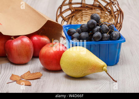 Giallo maturo pera, grappolo di uva in una scatola, un cesto di vimini, mele rosse con il sacchetto di carta e foglia secca sulla scrivania in legno. Un sano cibo biologico. Foto Stock