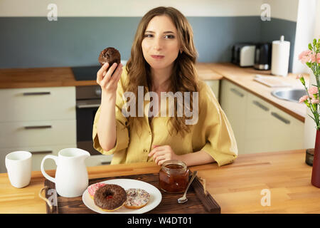Giovane bella ragazza è di avere la prima colazione a casa in cucina. Ella beve il suo caffè del mattino e si mangia un muffin al cioccolato. Foto Stock