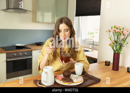 Giovane bella ragazza è di avere la prima colazione a casa in cucina. Ella beve il suo caffè del mattino e con grande piacere mangia la marmellata da una possibile. Foto Stock