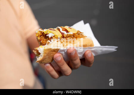 Un giovane uomo detiene un morso di hot dog nella sua mano. Snack nel parcheggio vicino al centro dello shopping. Foto Stock