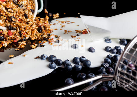 Yogurt versato dalla brocca di vetro, mirtilli versata da acciaio stainer e granola versata e diffusa dal recipiente di smalto miscelati insieme in un pasticcio su Foto Stock