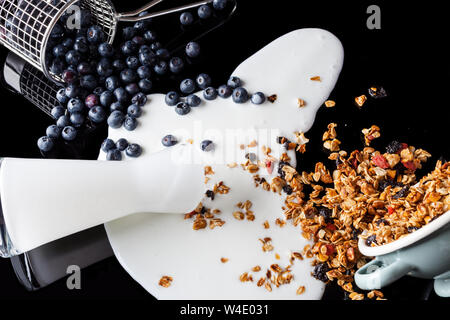 Yogurt versato dalla brocca di vetro, mirtilli versata da acciaio stainer e granola versata e diffusa dal recipiente di smalto miscelati insieme in un pasticcio su Foto Stock